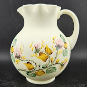 May include: Cream-colored pitcher with a ruffled rim and a curved handle. The pitcher is decorated with pink and yellow floral patterns and green leaves. A decorative piece for serving beverages.