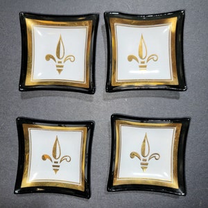 May include: Four square, decorative dishes with a white center, gold fleur-de-lis design, and gold trim. The dishes have a black border and a slightly curved shape. The dishes are likely for serving small items or as decorative accents.