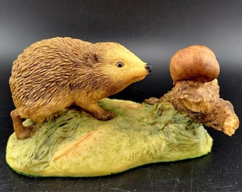 May include: A ceramic figurine of a brown hedgehog standing on a green base with a mushroom on a branch. The hedgehog is looking at the mushroom.