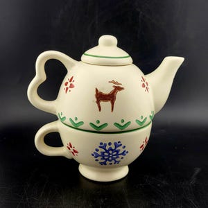 May include: Cream-colored ceramic teapot and teacup set. The teapot features a heart-shaped handle, a deer illustration, and red and green floral accents. The teacup has a snowflake design and a small handle.