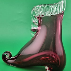 May include: A decorative glass vase shaped like a shoe, featuring a clear, ruffled top and a deep burgundy body. The shoe has a curled toe and a small, rounded heel. The vase is set against a green background.