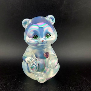 Pode incluir: Estatueta de urso iridescente com olhos verdes, nariz vermelho e um coração roxo no peito. O urso está sentado com as patas juntas e tem um pequeno objeto redondo nas patas. A estatueta tem um acabamento perolado.