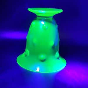 May include: A vivid green glass goblet with a flared base and a small, flat rim. The goblet features a series of raised, rounded bumps. The glass glows brightly against a dark blue background.