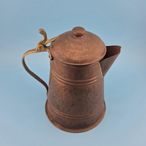 May include: A copper-colored metal pitcher with a tapered cylindrical body, a wide spout, and a domed lid. The pitcher features a curved brass-toned handle attached to a hinged mechanism on the lid, with horizontal embossed lines around the body.