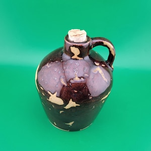 May include: A small, round, brown ceramic jug with a handle and a cork stopper. The jug has a dark brown glaze with lighter brown and beige accents. The jug is likely used for storing liquids.