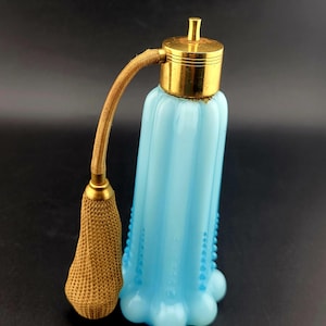 May include: A vintage light blue perfume bottle with a gold-colored metal top and sprayer. The bottle has a fluted design and a beaded pattern along the sides. The sprayer is connected by a gold-colored flexible tube.