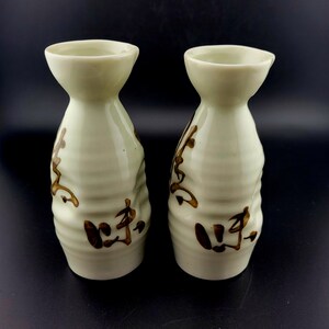 Puede incluir: Un juego de dos botellas de sake de cerámica con un esmalte verde claro y diseños pintados en marrón. Las botellas tienen un fondo redondeado y una boca ancha.