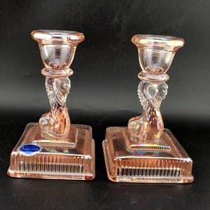 May include: Two pink glass candle holders. Each has a square base with a ribbed design, a decorative stem, and a cup-shaped top. The glass has an iridescent quality, reflecting light. A blue and white label is on the base.