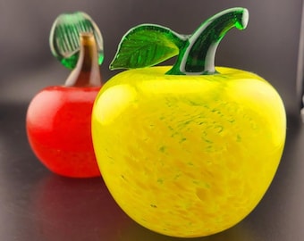 May include: Two glass apples, one red and one yellow with green leaves. The yellow apple has a swirl pattern.