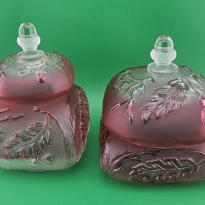 Op de afbeelding: Twee decoratieve glazen potten met deksels. De potten zijn diep roze van kleur met een matte afwerking. De deksels zijn helder en hebben een kleine, eikelvormige knop. De potten zijn versierd met een reliëf eikenblad- en eikelontwerp.