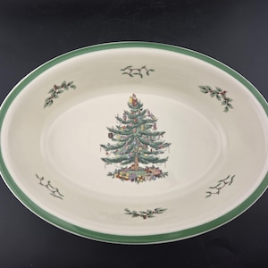 May include: An oval ceramic serving platter with a cream-colored base and a green rim. The center features a decorated Christmas tree with gifts underneath, while the border is accented with small sprigs of holly and mistletoe motifs.