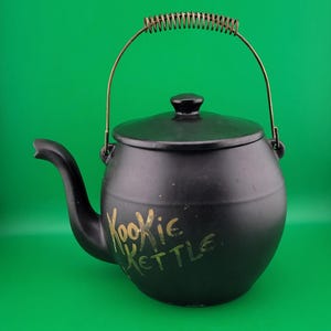 May include: Black ceramic kettle with a lid and a curved spout. The kettle has a gold-colored handle and the words "Kookie Kettle" in gold script on the side. The background is a solid green.
