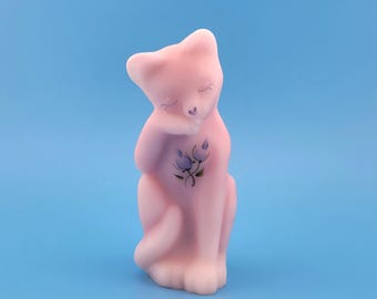 May include: A pink cat figurine with a light pink body and a white head and paws. The cat is sitting with its eyes closed and has a small purple flower design on its chest. The background is a solid light blue.