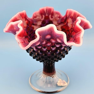 Puede incluir: Un cuenco de vidrio con borde ondulado, en tonos de rojo, morado y blanco. El cuenco tiene un patrón de clavos y una base de vidrio transparente. El borde tiene un diseño ondulado y con volantes.