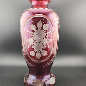 May include: A ruby red glass vase with a floral design etched into the glass. The vase has a round base and a narrow neck.