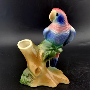 Puede incluir: Figura de loro de cerámica posada en una rama de árbol. El loro tiene plumas azules, rosas y rojas. La rama del árbol es marrón con hojas verdes. Es un objeto decorativo.