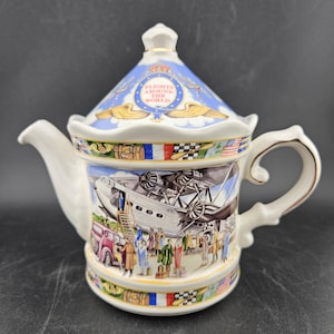 May include: A white ceramic teapot with a lid featuring a blue and gold design with the text "Flights Around The World." The body of the teapot depicts a vintage airplane and travelers. The handle and spout are white with gold trim.