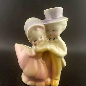 Puede incluir: Figura de cerámica vintage de una pareja abrazándose, con sombrero de copa y gorro. La figura está pintada en colores pastel, incluyendo rosa, amarillo y lavanda. La base es una maceta, y el diseño general es romántico.