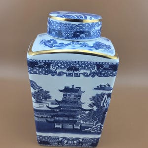 May include: A square ceramic ginger jar featuring a white base with intricate blue willow-style patterns depicting pagodas, trees, and bridges. The lid and the upper rim of the jar are accented with a thin, reflective gold metallic trim.
