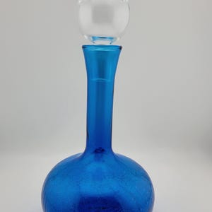 May include: A vibrant blue glass decanter with a clear, spherical stopper. The decanter has a round base and a long, slender neck. The glass has a textured, crackled appearance.
