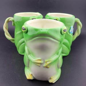 Puede incluir: Tres tazas de cerámica con forma de rana. Las tazas son principalmente verdes con vientres blancos y detalles amarillos en los pies. Las asas están diseñadas para parecerse a vides retorcidas. Cada taza tiene un diseño de cara de rana.