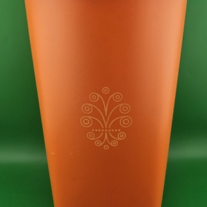 Puede incluir: Un vaso de plástico naranja con tapa. El vaso tiene una superficie texturizada y un diseño floral decorativo. El diseño está centrado en la parte delantera del vaso. El vaso está vacío y está sobre un fondo verde.