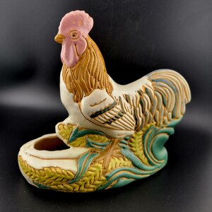 Puede incluir: Figura de gallo de cerámica con cresta y barbilla rosas, plumas marrones y blancas, y base verde y amarilla. El gallo tiene una pequeña abertura para guardar objetos. La figura es un artículo decorativo.