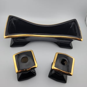 Puede incluir: Un centro de mesa de cerámica negra con ribete dorado y dos candelabros cuadrados a juego con ribete dorado. El cuenco tiene un diseño curvo y se apoya sobre dos pies. Los candelabros tienen una base cuadrada.