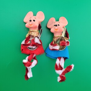 May include: Two whimsical mouse figurines with pink ears and faces, wearing red and blue skirts, and playing musical instruments. Their legs are made of red and white striped pipe cleaners. The background is a solid green.