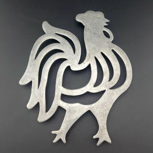 Puede incluir: Un salvamanteles de metal plateado con forma de gallo. El salvamanteles presenta un diseño recortado detallado de un gallo con cola curvada, cresta y patas con tres dedos. El salvamanteles está diseñado para proteger las superficies de los utensilios de cocina calientes.