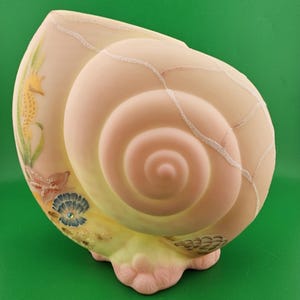May include: A ceramic seashell sculpture in shades of pink, yellow, and beige. The spiral interior is a soft pink, with a textured exterior and decorative accents of a seahorse, starfish, and seashell. The base is a light pink color.
