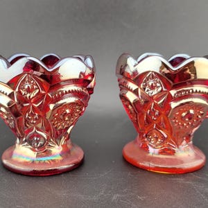 Op de afbeelding: Twee iriserende rode glazen eierdopjes met een gedetailleerd, gestructureerd patroon. De bekers hebben een geschulpte rand en een voet. Het glas heeft een glanzende, reflecterende kwaliteit en het ontwerp bevat ster- en bloemmotieven.