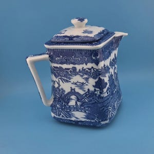 May include: A square-shaped ceramic teapot featuring a blue and white willow pattern. The design depicts a landscape with bridges, pagodas, and boats. It includes a matching lid with a small knob handle and a side handle for pouring.