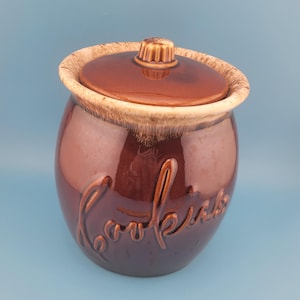 May include: A brown ceramic cookie jar with a lid and a decorative knob. The jar has the word "Cookies" embossed on the front. The rim and lid have a lighter brown, textured design.