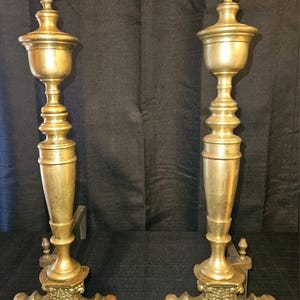 May include: A pair of matching brass fireplace andirons featuring tall, slender columns topped with flame-shaped finials. Each andiron rests on a wide, ornate base with intricate scrollwork and curved feet, providing a stable foundation.