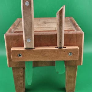 May include: A wooden butcher block with two knives inserted. The block is rectangular with a grid-like surface. The knives have wooden handles and green plastic blades. The block is supported by four legs and a wooden brace.
