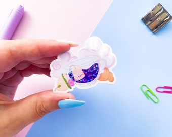 May include: A sticker of a teacup with a tea bag, a cloud, and a croissant. The teacup is filled with purple and blue glitter. The sticker is on a light blue surface.