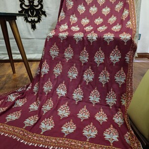 May include: Burgundy fabric with a repeating floral pattern in shades of peach, white, and blue. The fabric has a detailed border with a paisley design. The fabric is draped over a chair and table.
