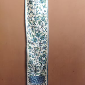 May include: A white tapestry with a blue floral design and a blue and white patterned vase at the bottom. The tapestry has a gold fringe at the bottom.