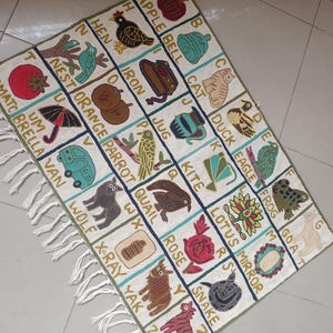 May include: A rectangular off-white textile wall hanging featuring an embroidered alphabet grid. Each square contains a letter, a corresponding illustration, and the printed word. The piece is finished with white fringe along the bottom edge.