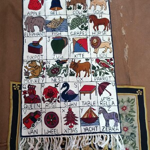 Peut inclure: Une tenture murale rectangulaire présentant une grille alphabétique brodée avec des illustrations pour chaque lettre de A à Z, incluant pomme, cloche, vache, chien, éléphant, poisson, raisin, cheval, encrier, cruche, cerf-volant, lion, singe, nid, bœuf, perroquet, reine, rose, cygne, table, parapluie, fourgon, roue, sapin, yacht et zèbre, avec une frange blanche en bas.