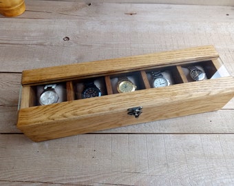 May include: A wooden watch box with a clear glass top. The box has five compartments for storing watches. The box is closed and the watches are visible through the glass top.