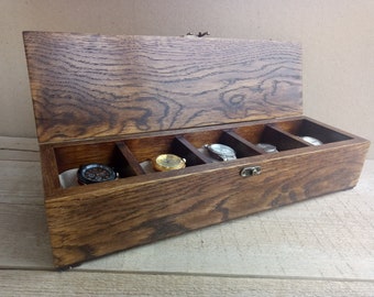 May include: A wooden watch box with a hinged lid, showcasing a rich, dark brown wood grain. The box is divided into five compartments, each holding a wristwatch. The watches have various band and face designs, including gold and silver tones.