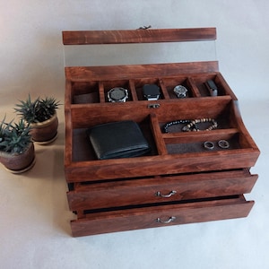May include: A wooden watch and accessories organizer with a clear glass top. The organizer has multiple compartments for watches, a wallet, bracelets, and rings. The wood has a rich, reddish-brown stain. Two drawers are visible at the bottom.