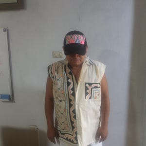 May include: A man wearing a white sleeveless shirt with a black and colorful geometric pattern. He is also wearing a black baseball cap with a colorful geometric pattern on the front. The man is wearing white pants.