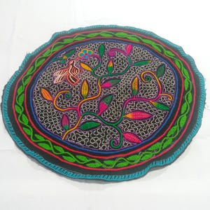Puede incluir: Textil bordado negro de forma redonda con un diseño floral colorido. El diseño central presenta un pájaro y enredaderas con hojas en tonos rosa, amarillo y verde. El borde tiene un patrón azul, rojo y verde.