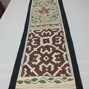 May include: A long, rectangular table runner with a black border and intricate patterns. The central design features brown and white geometric shapes, a circular motif with a bird, and green leaf accents. The runner is displayed on a white surface.