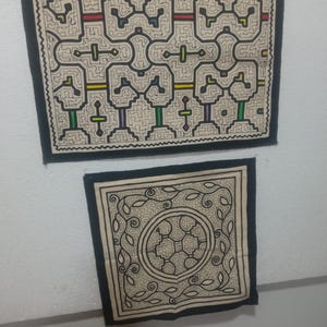 Op de afbeelding: Twee ingelijste textielkunstwerken. Het grotere stuk heeft een complex geometrisch ontwerp in zwart, beige en accenten van rood, geel, groen en paars. Het kleinere stuk heeft een centraal cirkelvormig ontwerp met omringende bladpatronen.