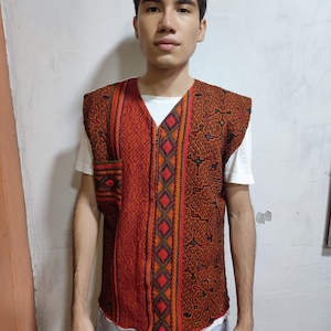 Könnte beinhalten: Eine rote, ärmellose Weste mit einem geometrischen Muster in Schwarz, Orange und Grün. Die Weste hat einen Reißverschluss vorne und eine Tasche auf der linken Seite.