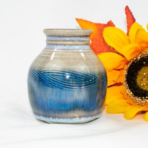 May include: A small, hand-thrown ceramic vase with a blue and white glaze. The vase has a rounded body and a slightly flared lip. It is decorated with a pattern of blue lines.